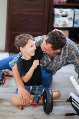 Baba oğul dört tekerli bisikleti tamir ediyor, bir adam çocuğa nasıl delineceğini gösteriyor, gülümsüyor ve birlikte eğleniyorlar, baba sevgisi
