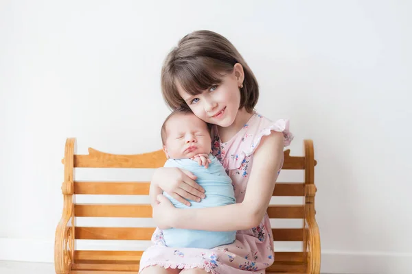 A girl with a short haircut in a pink dress holds a newborn in a blue cocoon, children in a white room, an older sister and a younger brother, a happy sister