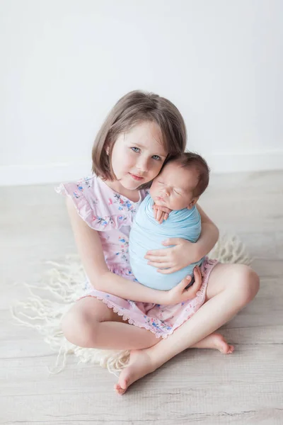 A girl with a short haircut in a pink dress holds a newborn in a blue cocoon, children in a white room, an older sister and a younger brother, a happy sister