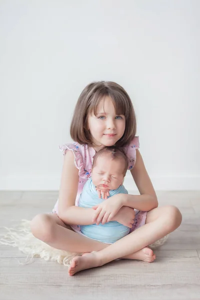 A girl with a short haircut in a pink dress holds a newborn in a blue cocoon, children in a white room, an older sister and a younger brother, a happy sister
