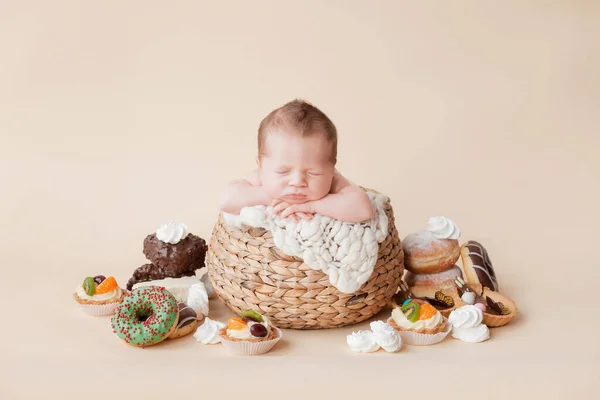 a little newborn baby sleeps in a straw basket on a beige background, there are many donuts, cakes and sweets around him