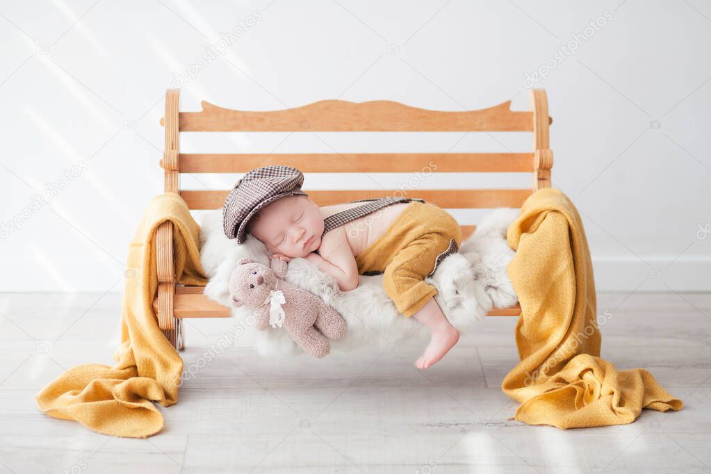 a newborn baby in yellow pants and a brown hooligan's cap sleeps on a wooden bench in a light room, hugs a toy bear