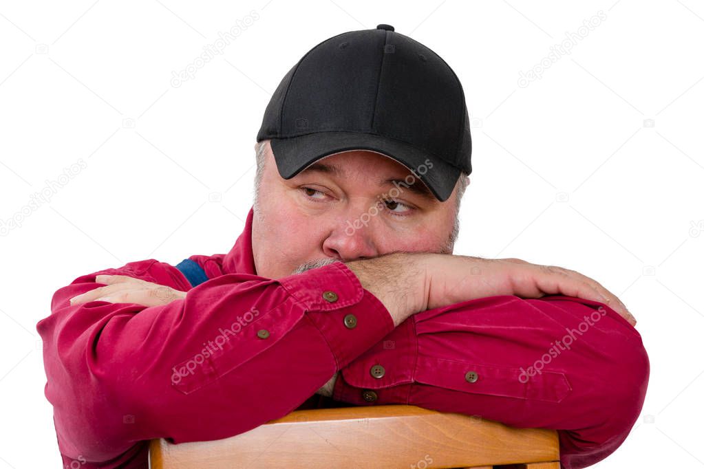 Thoughtful farmer or worker in a peaked cap leaning on his arms on the back of a wooden chair looking to the side with a watchful pensive expression isolated on white