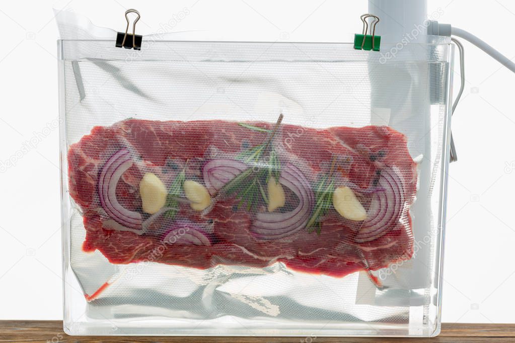 Portion of flat iron beef steak sous-vide cooking suspended in a clear plastic bag in hot water with a spirg of fresh rosemary and onions to flavor