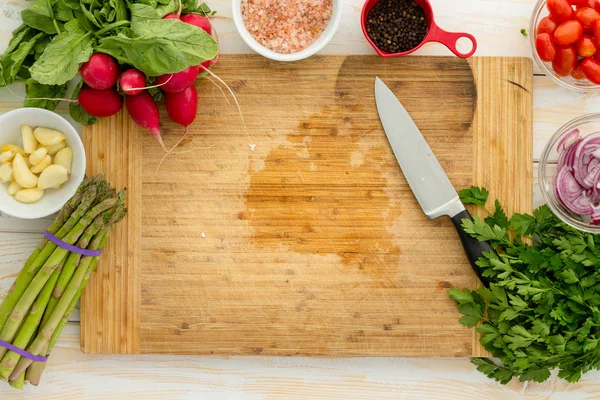 Overhead view of wooden cutting board surrounded by assorted vegetables including onion and asparagus with knife