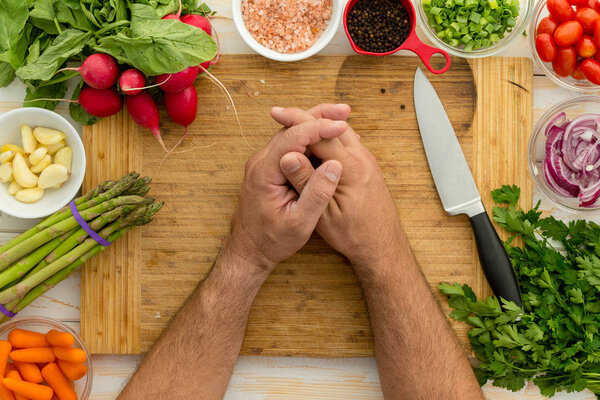 Man resting hands on wooden cutting board bordered by multiple vegetables including green pepper and carrots next to small bowls of spices