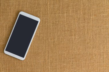 Hafif kahverengi çapraz iplik oluşan yüzey üzerinde oturan bir beyaz akıllı telefon. Kopya alanı içerir.