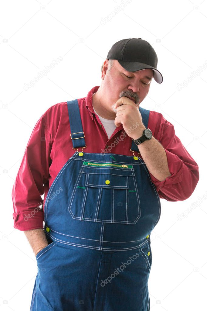 Big man in denim bib overalls and cap standing thinking looking down at the floor with his hand to his chin isolated on white