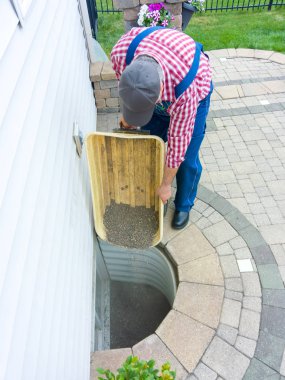 Man replacing the old pebbles in an egress window in the basement of his house during spring maintenance emptying the stones from a wheelbarrow clipart