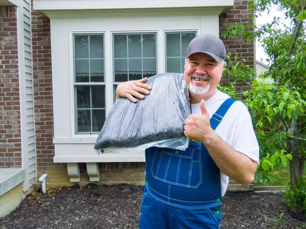 Happy strong gardener or or garden services worker happy to be mulching your garden giving a thumbs up with a beaming smile as he carries a bag of mulch