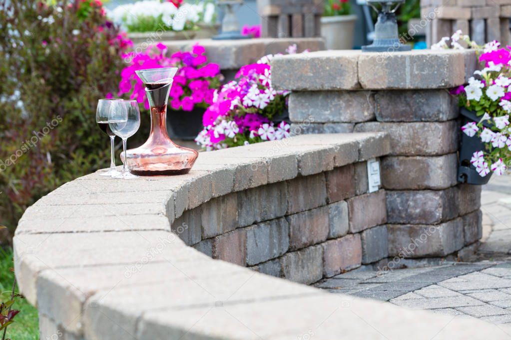 Decanter of red wine aerating on a curved brick patio wall with two wineglasses in front of colorful potted summer flowers with copy space
