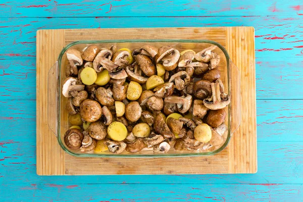 Glass oven dished filled with mushrooms and diced potatoes ready to be roasted as a healthy accompaniment to a meal on a bamboo board over blue viewed from above