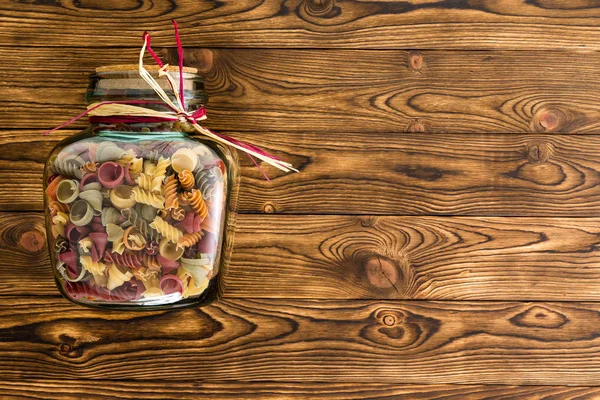 Clear glass storage jar with macaroni fall pasta in a mix of shapes and colors laid on its side on a wooden background with copy space