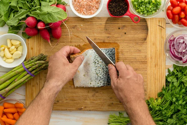 男子准备一个新鲜的三文鱼鱼片烹饪清洁皮肤用一把锋利的刀在板上被新鲜的沙拉成分包围 关闭头顶上的手的看法 — 图库照片