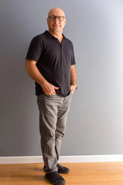 Full length portrait of adult middle-aged bold man in glasses, wearing black polo and grey jeans, standing in half turn with his fingers in pockets and looking at camera