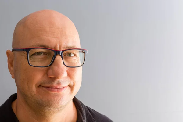 Close-up bust front portrait of smiling bold middle-aged man in black-rimmed glasses and black shirt, looking at camera with a friendly smile. Plain grey wall background with copy space