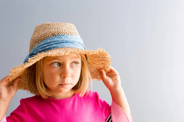 若い可愛いピンクのドレスと両手で彼女の帽子を押しながら普通の灰色の壁の背景にコピー スペースの側に離れている農村の麦わら帽子の女の子 — ストック写真