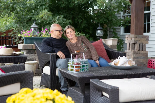 Adult couple sitting close together on black outdoor couch at beautiful patio, and enjoying their time. Bold man in glasses hugging woman in warm brown sweater