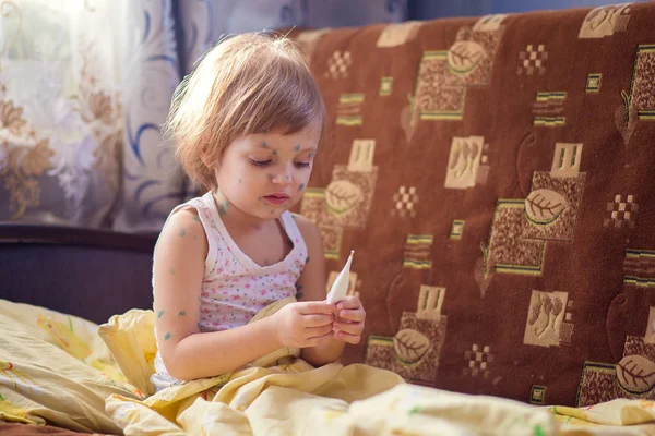 The child sick chicken pox sits in a bed and holds the thermometer in hand