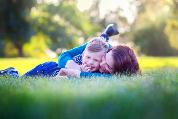 Young mother with the son lie on a grass in the park and joyfully embrace — Stock Photo, Image