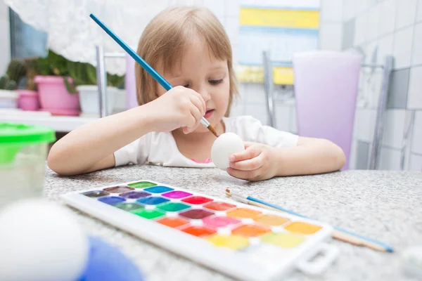 The little girl at home in kitchen paints Easter eggs with a brush and watercolor paints