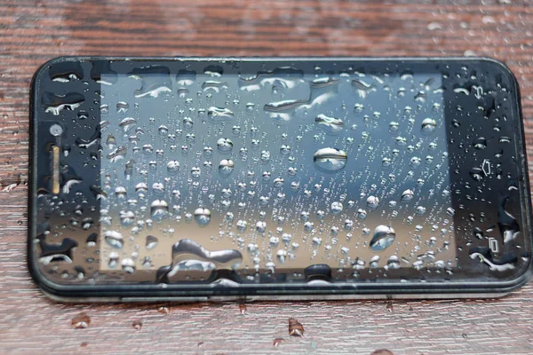 Primer plano del teléfono inteligente negro que se llena con gotas que se encuentra en una mesa de madera marrón — Foto de Stock