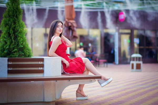 The Armenian girl in a red dress sits on a shop in the summer city near cafe. Air irrigation by water in the hot city