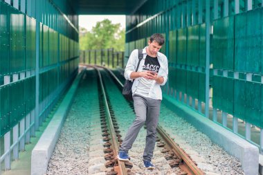 Raylar üzerinde bir sırt çantası maliyeti ile beyaz gömlek li genç adam ve telefonda bir şey arar