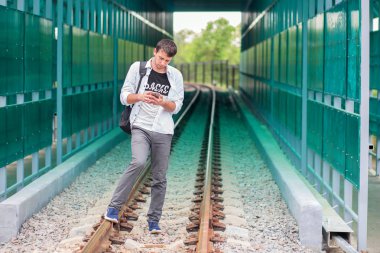 Raylar üzerinde bir sırt çantası maliyeti ile beyaz gömlek li genç adam ve telefonda bir şey arar