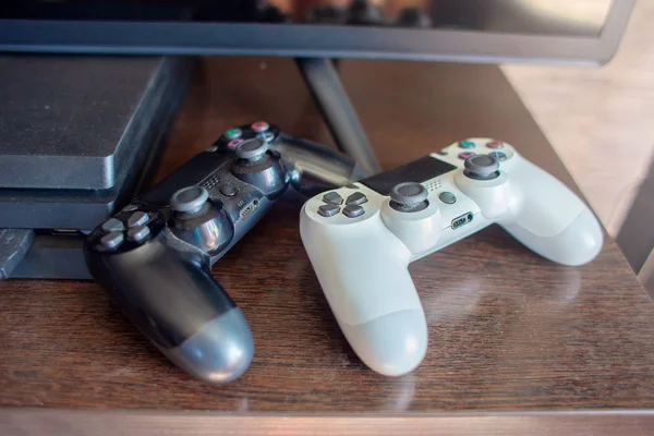 Dos joysticks de la consola de juegos - chery y blanco - se encuentran en una mesa cerca del televisor y la consola de juegos — Foto de Stock