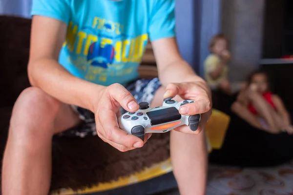 The young guy in a blue t-shirt stays at home on a sofa, holds the joystick in hand and plays on the game console — Stock Photo, Image