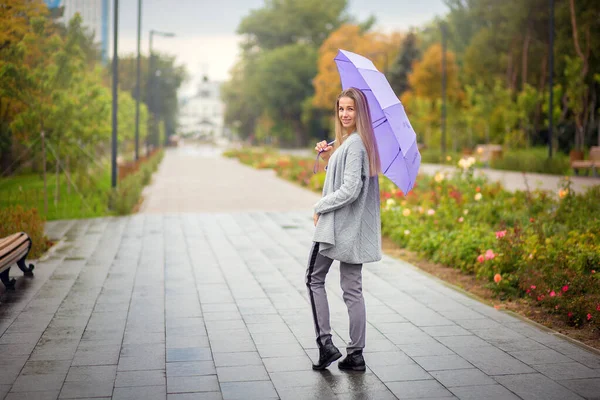 Una donna in pantaloni grigi con un ombrello lilla passeggia nel parco autunnale — Foto Stock