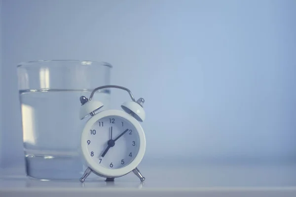 White alarm clock and glass of water on blue tones