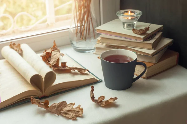 autumn still life. books, leaves, cuo of tea and dry tiny flowers in vase red candlestick with a burning candle on window. Concept of autumn reading time and romantic, Warm, cozy window seat  opened book, light through shutters, rustic style home dec