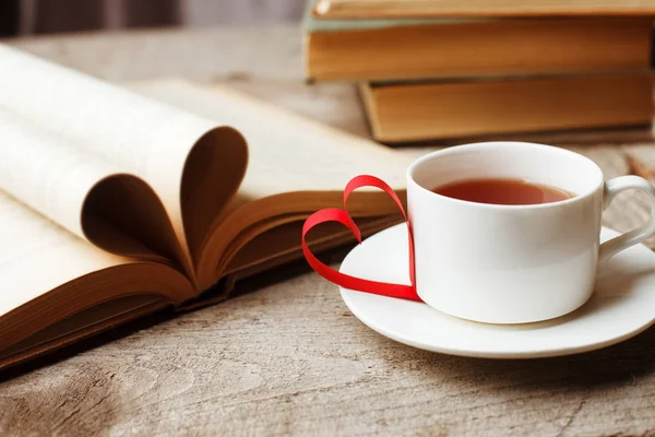 Love of books, reading. Stack of books on the wooden table. Open book with curled leaves in the shape of a heart, cup of tea. Library, education, leisure time concept.