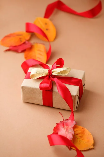 Autumn creative holiday present. Handmade paper gift box with satin bow, foliage dried leaves on beige background. Thanksgiving day, fall background. Copy space, stylish card