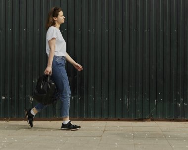 Çitboyunca yürüyen bir kadın. Kot pantolon ve t-shirt giyen ve kentsel arka planda yürüyen sırt çantası tutan genç yan görünümü.