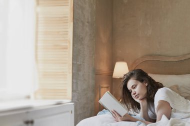 Genç bir kadın yatağa uzanıp kitap okuyor.