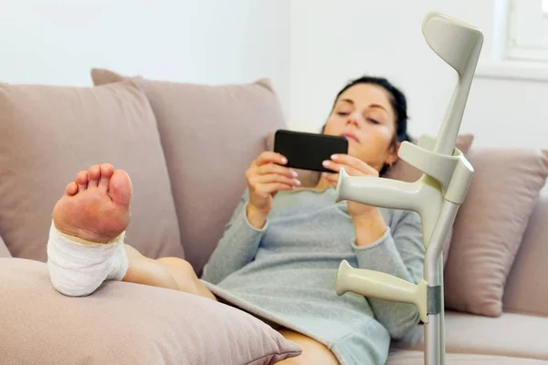 Mulher Com Muletas Deitada Sofá Usando Smartphone — Fotografia de Stock