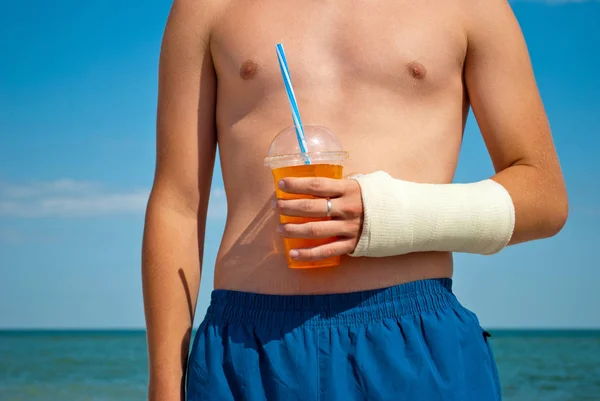 Gypsum fracture on a man\'s hand, cocktail juice orange, sand close-up against the background of the sea and the sky clouds, broken arm limb, illness recovery, the guy in blue shorts, summer sun
