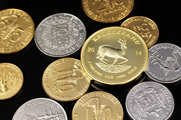 A macro image of an assortment of West African Franc coins and a gold South African one ounce Krugerrand on a reflective black background