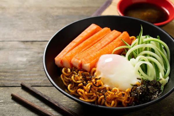 Spicy instant noodle with crab stick and onsen egg and cucumber in black bowl on wooden table.
