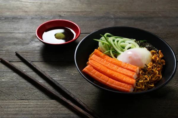 Spicy instant noodle with crab stick and onsen egg and cucumber in black bowl on wooden table.