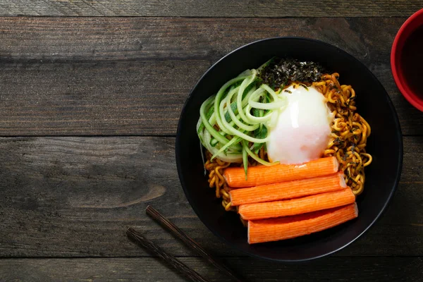 Spicy instant noodle with crab stick and onsen egg and cucumber in black bowl on wooden table.