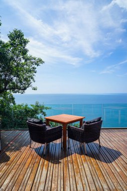 Tablo ve rattan açık koltuk ayarı ahşap güverte ve cam balkon panoramik Andaman okyanus deniz görünümü, yeşil ağaç ve mavi gökyüzü arka plan ile güneşli, Phuket, Tayland