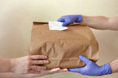 Mavi lateks eldivenli erkek eller kadın ellerine kese kağıdı veriyor. Eldivenli erkek ellerinin yakın çekimi beyaz etiketli kahverengi el işi kağıt torba tutuyor. Teslimat hizmeti konsepti. Boşluğu kopyala.