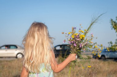 Vahşi çiçekler bir buket ile güneşli bir günde araba ile yolun yanında bir alanda küçük sevimli sarışın kız. Arka görünüm.