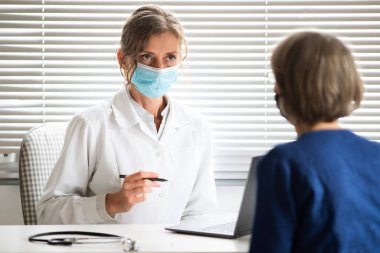 Kadın doktor, tıbbi maske takan bir hastaya danışıyor.