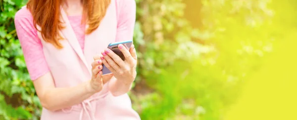 Banner Beautiful young girl sitting on a wooden bench in the open Looking at the mobile phone. Women\'s hands Sunny day green bushes lifestyle Selective focus copy space