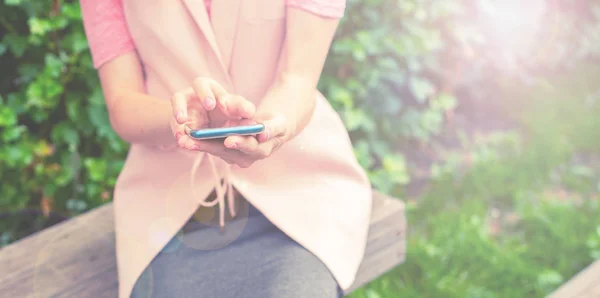Banner Beautiful young girl sitting on a wooden bench in the open Looking at the mobile phone. Women\'s hands Sunny day green bushes lifestyle Selective focus copy space